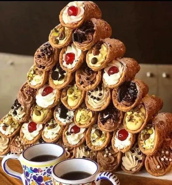 A pyramid of cannolis stacked in front of two cups of coffee