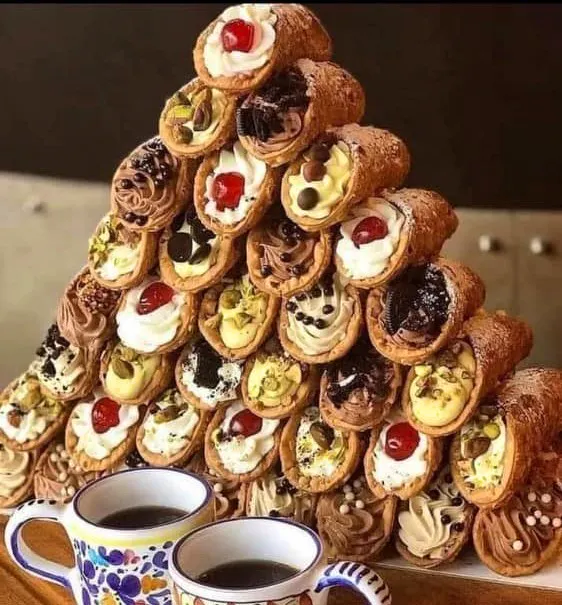 A pyramid of cannolis stacked in front of two cups of coffee